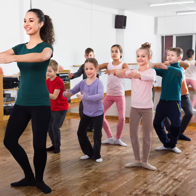 A group of ballet students stand behind the ballet instructor. They are copying the ballet move she is demonstrating.