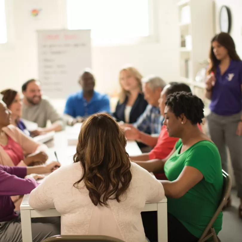 Group of people sit at a table looking at a booklet and paying attention to a teacher who is talking near a whiteboard