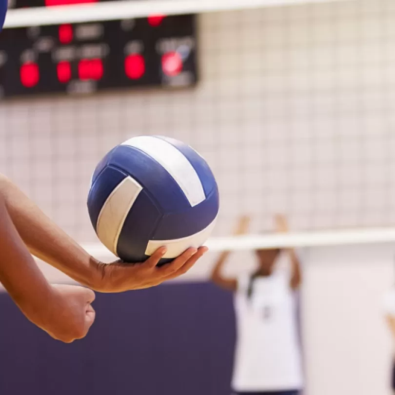 A unrecognizable person prepares to serve a volleyball during a game inside a gym.