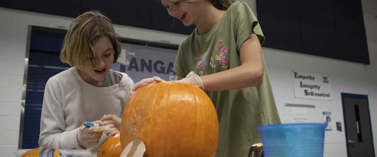 Two pre-teen girls work together to carve a pumpkin, both are laughing.