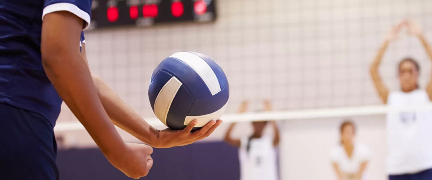 A unrecognizable person prepares to serve a volleyball during a game inside a gym.